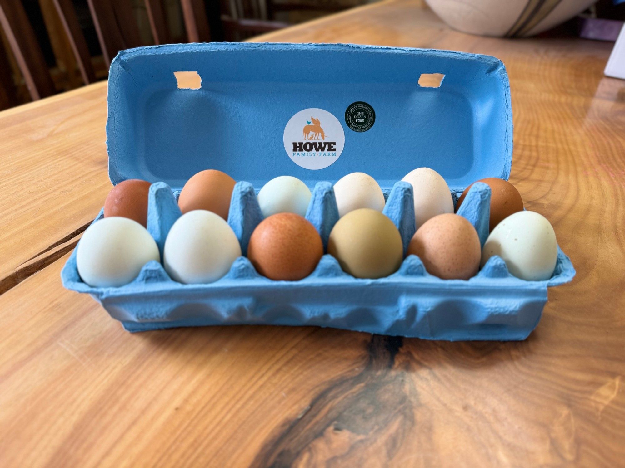 An open blue Howe Family Farm carton holding a dozen eggs in shades of brown, cream, pale blue and olive